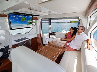 Two people seated on the salon settee of a Pursuit OS 445 with the television on.