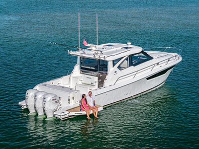 Two people seated on the swim platform of a Pursuit OS 445 at rest, feet in the water beside the outboards.