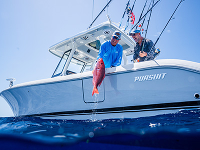 Two men lean over the side of a Pursuit boat holding up a freshly caught red snapper, with fishing rods mounted in rocket launchers overhead.