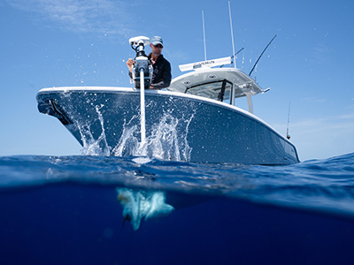 Split-level photo of a man operating a bow-mounted trolling motor on a boat, with water splashing at the surface and a ray visible swimming underwater below.