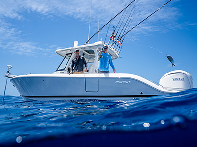 Two men on a Pursuit boat with one reeling in a fish that's leaping out of the water on the line.