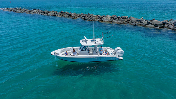 Pursuit center console boat anchored near a rock jetty in calm turquoise water, with two adults and two children standing in the cockpit.