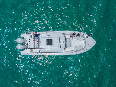 Aerial overhead view of a Pursuit center console boat drifting on sparkling green water, with three people on the aft deck and one holding a fishing rod.