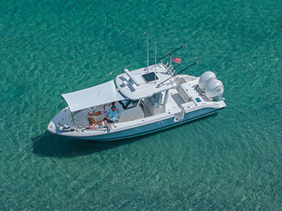 Pursuit center console boat anchored in clear turquoise water with a shade canopy extended over the cockpit, where two people are seated, and an anchor line visible off the bow.