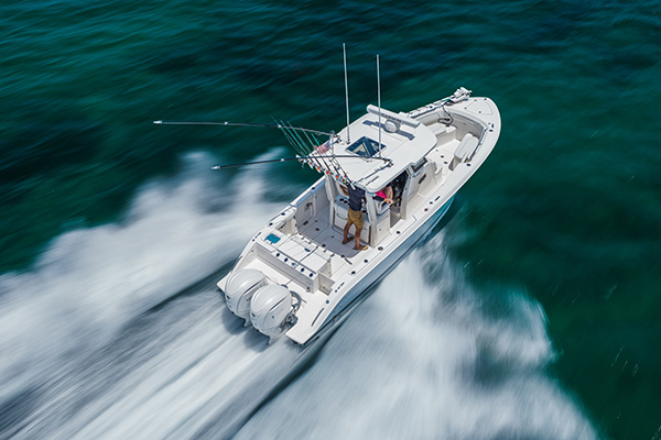 Pursuit center console boat turning at speed on teal-green water, with a man standing at the helm and a woman seated beside him as the twin outboard engines throw up a white wake.