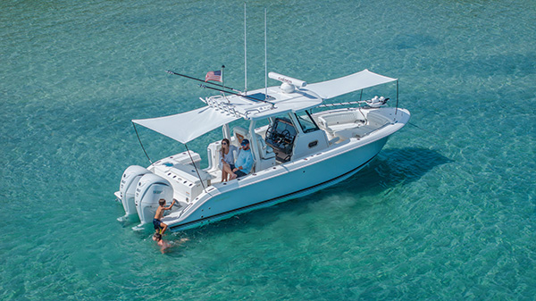 Pursuit center console boat anchored in shallow turquoise water with its sun shade extended, a man and woman seated in the cockpit, and two children climbing and swimming near the stern.