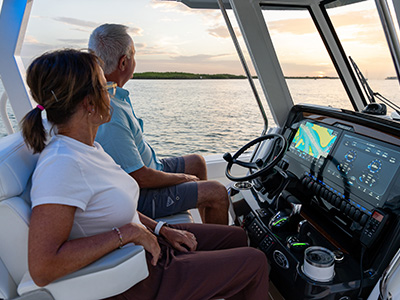 Man and woman seated at the helm of a Pursuit boat at sunset, viewed from behind, with the multifunction display showing a chart and engine data.