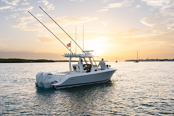 Pursuit center console boat idling at sunset with three people aboard, a marina and sailboats visible in the background.