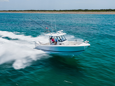 Pursuit center console boat running parallel to a sandy shoreline, with three people aboard and fishing rods mounted upright on the hardtop.
