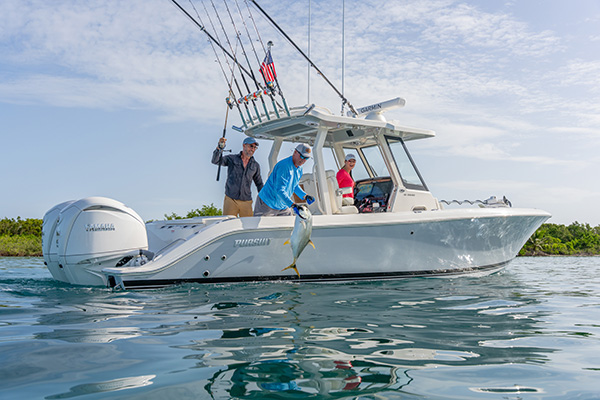 Man lifting a freshly caught jack fish from the water at the stern of a Pursuit boat, with two people watching and laughing nearby.