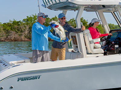 Two men holding up a freshly caught jack fish and taking a selfie on a Pursuit boat, with a woman at the helm in the background.