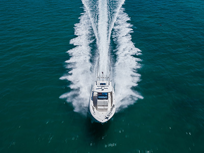 Aerial view of a Pursuit center console boat running directly toward the camera, its wake spraying symmetrically on both sides and several fishing rods standing upright near the bow.