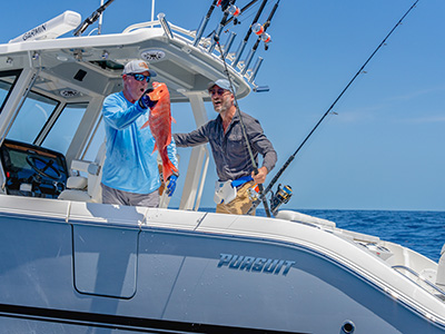 Close-up of two men on a Pursuit boat, one holding a freshly caught red snapper near the camera, with the Pursuit hull lettering visible behind them.