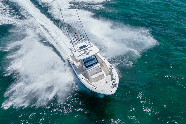 Pursuit center console boat crossing through a large wake with spray breaking over the bow, cockpit empty, fishing rods mounted upright on the hardtop.