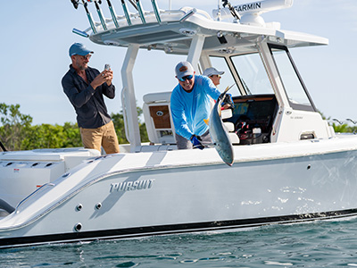 Man releasing an amberjack over the side of a Pursuit boat while another man photographs the moment on his phone, with a mangrove shoreline in the background.