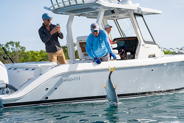 Man releasing an amberjack that splashes into the water beside a Pursuit boat, while another man photographs the moment and a woman looks on from the helm.