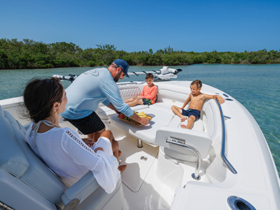 Man serving a fruit platter to two boys seated on the bow cushions of a boat, with a woman seated nearby and a mangrove-lined channel in the background.