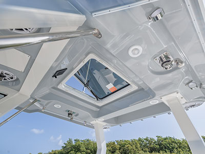 Underside view looking up at a Pursuit boat's hardtop, showing an open overhead escape hatch and speaker mounts against a blue sky and mangrove treeline.