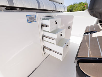 Close-up of an open storage drawer unit built into the side console of a Pursuit boat, showing three pull-out bins beside a teak-accented bench seat.