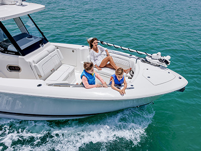 Aerial view of a Pursuit S 288 center console boat running at speed, with a woman lounging on the bow cushions and two boys in life jackets leaning over the bow rail.