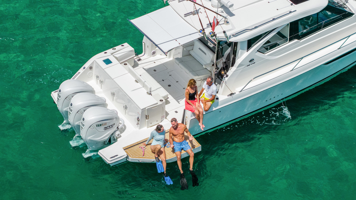 Two people in snorkel fins seated on the swim platform of a Pursuit OS 445 over green water.