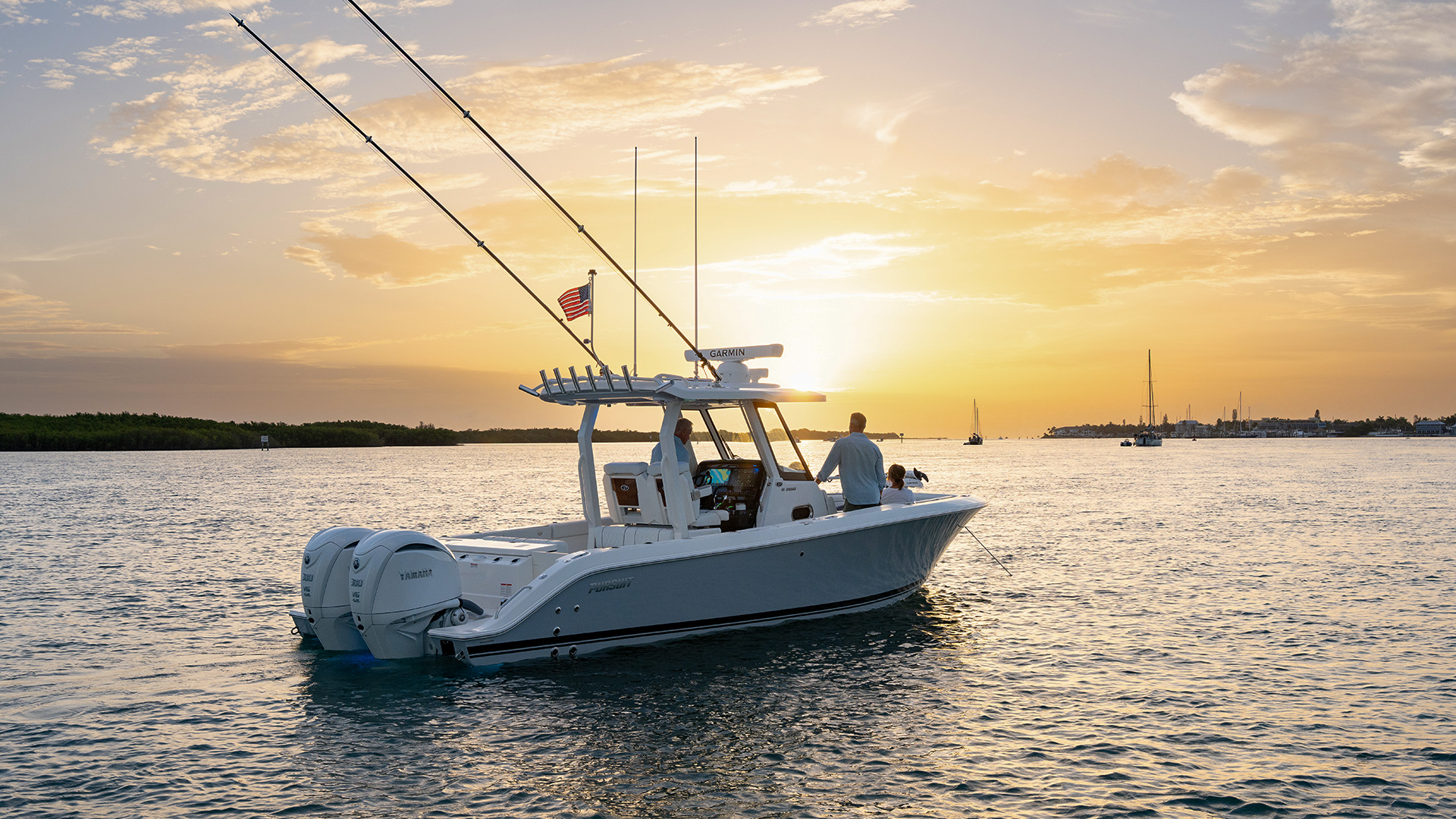 Pursuit center console boat idling at sunset with three people aboard, a marina and sailboats visible in the background.