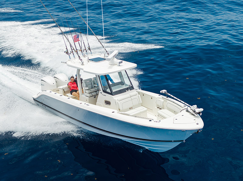 Pursuit sport boat speeding across open blue water, viewed from above, with fishing rods, twin outboard engines, and passengers aboard.
